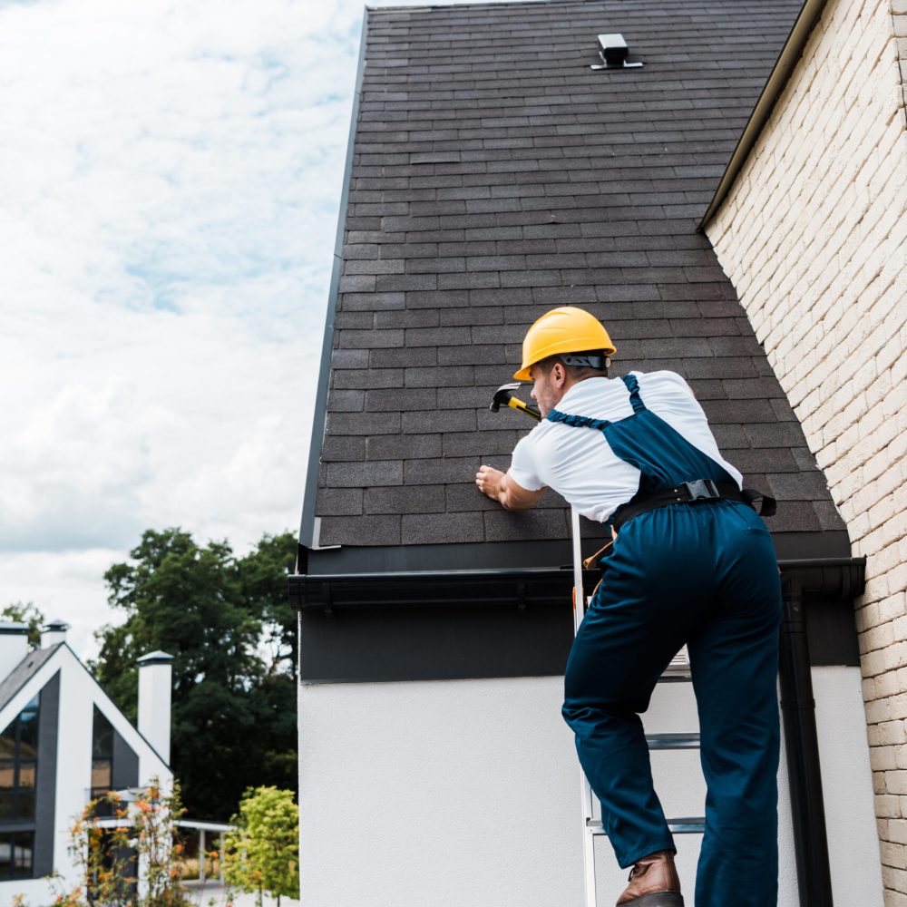 repairman in uniform and helmet repairing roof while standing on ladder